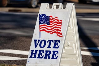 A picture of a small polling place sign instructing voters to “Vote Here.”