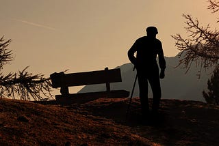 An old man with a cane stands by a bench with a mountain view.