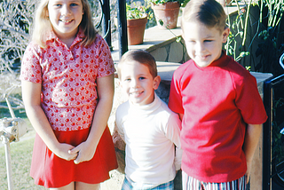 A young white girl about eight, a little guy in the middle, probably three years old and another brother, on the far right, who’s about five or six. We’re all wearing patriotic, if not oddly cobbled together, outfits. Circa 1970.