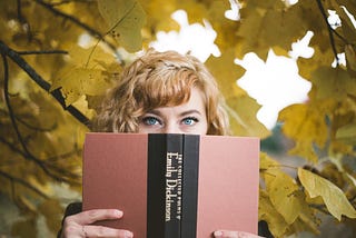 A woman staring straight at the camera, the lower half of her face hidden by an open book, which is “The Collected Poems of Emily Dickinson”