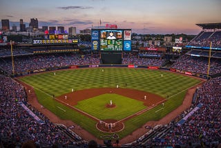 Three level baseball stadium filled with fans with players in positions on the field. Green grass, brown infield and huge lit-up screen with player stats at sundown with dark clouds along the skyline.