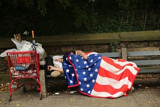 A houseless man sleeps on a bench next to a shopping cart full of his belongings, draped in an American flag blanket.