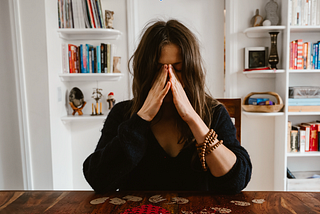 Caucasion appearing woman sitting at a wooden table in a black sweater holding her hands in prayer shape over her face looking distressed. She sits in front of a bookshelf with colorful books.