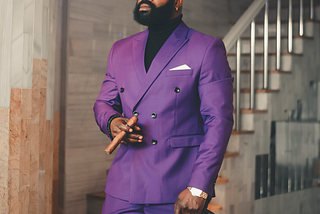 A black man like me, dressed to the nines in a purple tailored suit, strolls out of a lavish home with a cigar in hand.