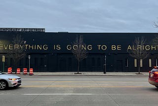 On the side of a building, the words “EVERYTHING IS GOING TO BE ALRIGHT”.