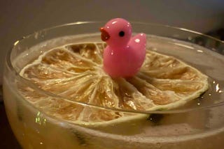 A light yellow cocktail in a clear glass topped with a dried citrus wheel, with a small pink plastic duck figurine sitting on the citrus slice.
