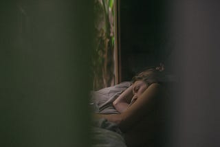 Photograph of a white woman lying on a bed in a dark room. He eyes are open, she is laying on her side. The photo is dark and the edges are blurred.