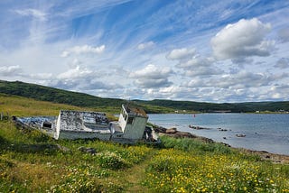 Old wooden boat with peeling paint on a grassy hill overlooking a bay on a sunny day.