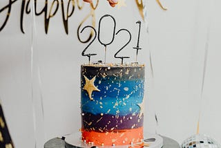 A colorful tall cake with “2021” on top if it and the words “Happy New Year” above the cake.