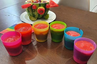 Six cups full of smoothies sit in a semi-circle in front of a hollowed out watermelon, carved in the shape of a pig, full of fruit.