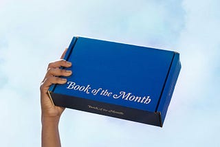 A hand holding a bright blue cardboard box, about the size of a book, up to the sky.