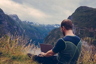 A digital nomad sitting on the grass, on top of a mountain working on his laptop