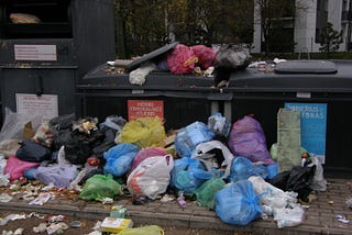 Residential trash bin area with huge bags & smaller debris pile dall around the bins as well as on top of them.