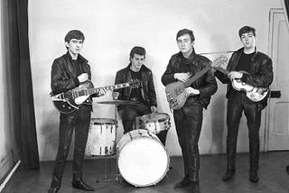 Four young men wearing leather jackets and holding their respective instruments. From left to right, George Harrison, Pete Best, John Lennon and Paul McCartney.