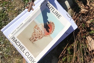 Book cover on dried grass, Outline by Rachel Cusk, with shadow of dinosaur tail over book.