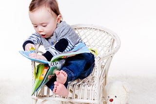 A young child, sitting in a chair, next to his white teddy bear, reading a picture book to himself.