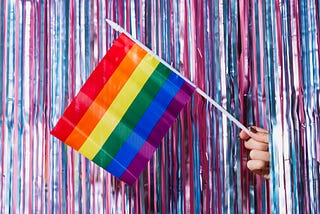 A small pride flag held from beyond a shiny curtain