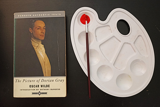 A paperback copy of The Picture of Dorian Gray on a dark background next to a palette with a paintbrush dipped in red paint.