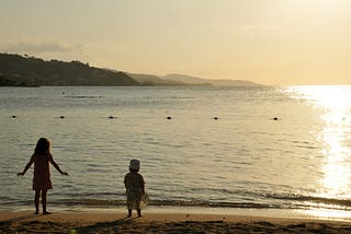 Two young children walk on the shoreline at sunset.