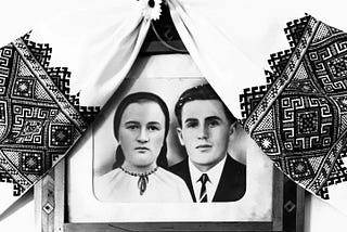 A black and white framed photograph of two people, draped by an embroidered hanging.