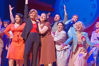 The author wearing a black suit with a red blouse raising one finger high, surrounded by a group of actors smiling out at the audience.