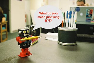 Toy robot holding a pencil and saying “What do you mean just wing it?”