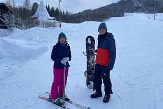 A male and female Asian couple in winter sports gear on a ski slope. The female is on skis, holding ski poles and the male is holding a snowboard. The female is wearing a green beanie, grey neckwarmer, dark blue ski jacket, pink salopettes and a pair of white gloves. The male is wearing a blue beanie, a two-coloured ski jacket where the top half is dark blue and the bottom half is orange with black salopettes, snowboard boots and gloves.