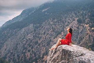 a girl in a red dress on top of a cliff admiring the view of a mountain gorge