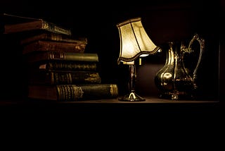 A crooked lamp illuminates a stack of books and a metal pitcher with low light