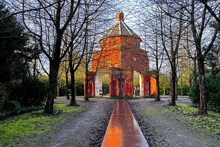 A soggy winter’s day in a public park, bare trees line a slick walkway that leads to a red brick rotunda, the lower level has high opan arches and tiny figures are walking through