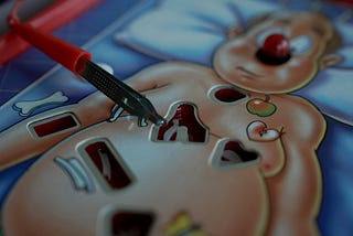 Close-up of the board game Operation: a metal tweezers hovers over the cartoon patient’s cut-out cavity while his red-nosed face watches anxiously.