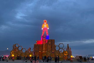 The man at dusk / moonrise, lit up in neon atop of beehive structure with gather clouds in the background