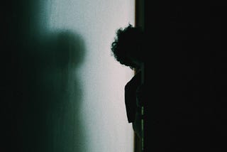 Photo Credit: Ammar Sabaa/Unsplash. A barely lit hallway and completely dark room. A boy shyly peeks around the corner of the dimly lit hall into the dark room.
