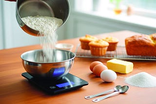 Digital kitchen scale for baking on a countertop with flour, eggs, and butter, surrounded by fresh baked cupcakes and bread in a modern kitchen.