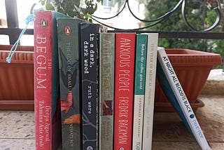 8 books are lined up in a standing position. From left to right, the books are: The Begum, The God of Small Things, In a Dark Dark Wood, A Wedding in December, Anxious People, Before the Coffee Gets Cold, The Year of Magical Thinking, At Night All Blood is Black.