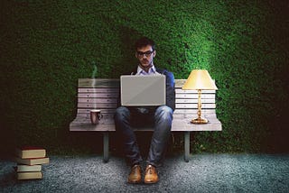 person dressed in men’s business casual with blue jeans, sitting on a bench against a backdrop of a dark green wall covered in artificial moss. person is typing on a laptop and has an electric lamp, a mug of a steaming drink, and a stack of books.