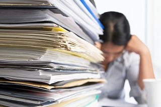 Person sitting at a desk behind a large stack of papers with head in hands.