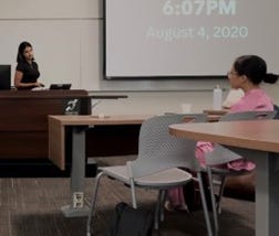 Sarah in black speaking in front of a podium to a class of students.