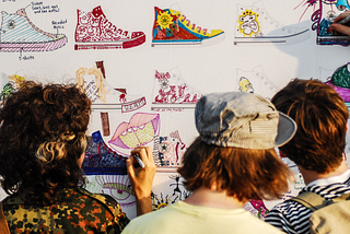 A color photo of three young New Yorkers stand before large piece of white poster paper covered with images of Converse All-Star hightop shoes, which they are applying their own designs too with different colored markers.