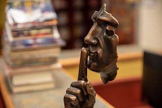 A bronze statue of a face with the hand up and one index finger over the lips.