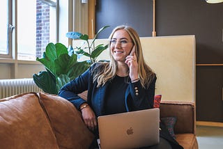 Blonde haired woman wearing glasses smiling while holding her phone to her ear and sitting on a leather couch with her laptop on her lap.