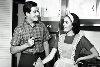 A black and white photo of a young married couple in the kitchen, with the woman wearing an apron and holding an iron.