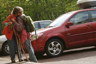 The author and her mother stand by a red vehicle after coming back from a long hike. They are dressed in hiking attire and carry walking sticks and backpacks. They share a hug of relief, happy to be done with a challenging journey.