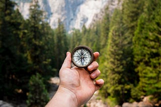 A hand holding up a navigational compass in front of a forest and mountains scene