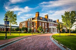 A color photograph showing the gravel drive across leading to Mason Hall, Johns Hopkins University, Baltimore MD, in the background. The green grass and trees of the quad are in the foreground to the left and right of the drive.