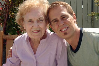 Elderly woman looks into the camera — young man has his arm around her smiling.