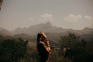Woman with her hands by her chin, contemplative. She’s in the center of the photo, side profile. There are mountains behind her.