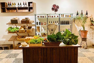 A local greengrocery cozy store with vegetables such as lettuce and tomatoes in the front area and in the back there are wine bottles, a vase with a plant, and other grocery products.