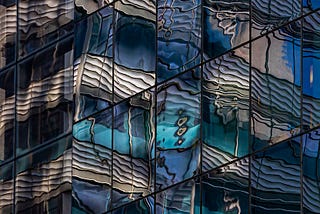 “Blue and white concrete building”, photograph by Robert Ullman (source: https://artist.scop.io/image/blue-and-white-concrete-building-73) — a building façade of square glass panes reflecting a distorted image of a concrete building in shades of dark blue, aqua, and off-white.