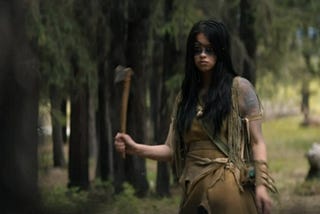 A young indigenous woman with long black hair and wearing buckskin stands in a forest in daylight. She has black face paint across her eyes and holds a tomahawk in her right hand.